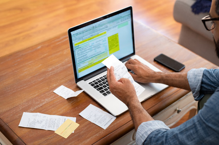Hands of businessman analyzing invoice on laptop while checking bills. Rear view of latin man checking invoice while matching them on computer. Closeup of man hands calculating financial expenses while working on laptop.の写真素材