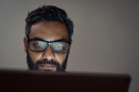 Mature business man wearing eyeglasses sitting at desk and using laptop in a dark office. Middle eastern man working on his computer while sitting at his working place at night time. Overworked latin man with specs at workplace in office in front of laptop with copy space.の写真素材
