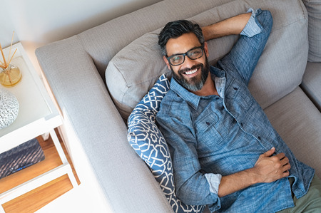 Happy mature man with eyewear and beard relaxing on sofa at home. Handsome latin man with hands behind head sitting on couch in living room. High angle view of cheerful indian man in casual lying on gray couch and looking at camera.の写真素材