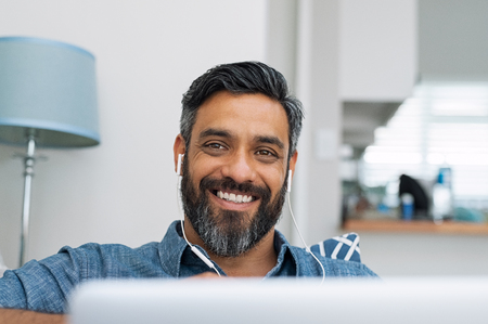 Portrait of happy man using laptop with earphones while lying on couch. Casual man in video call lying on sofa while listening to music with computer. Mature middle eastern man with earbuds looking at camera with a big smile.