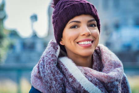 Closeup face of a young happy african woman enjoying winter wearing scarf and cap. Smiling brazilian girl in a colorful shawl looking at camera in a cold day. Multiethnic woman with knitted bordeaux hat and woolen scarf.の写真素材