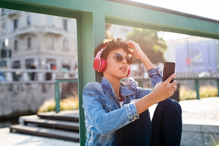 Young african woman listening to music with headphones on street. Portrait of brazilian girl listening music online with wireless headphone from a smartphone. Urban girl enjoy songs sitting on the ground in a summer sunny day.の写真素材