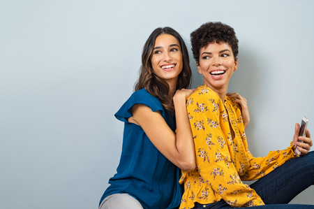 Two smiling young women sitting together and looking away. Happy multiethnic girls having fun isolated on gray background. Beautiful stylish friends laughing while sitting on bench with copy space.の写真素材