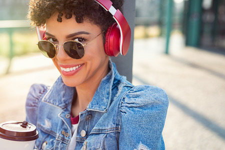 Portrait of a cheerful stylish woman listening music with red headphones and looking at camera. African young woman holding coffee while taking a break. Closeup face of brazilian girl listening music with vintage sunglasses.の写真素材