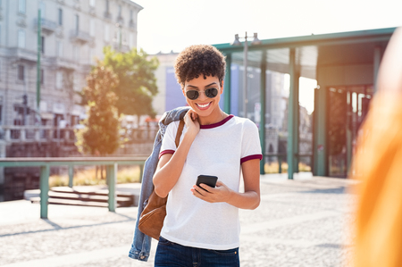 Smiling african woman using mobile phone while walking on street. Happy tourist searching for map directions on smartphone. Stylish brazilian girl wearing sunglasses looking at cellphone while travelling.の写真素材
