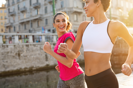 Two sporty women jogging in city. Multiethnic girls running on street at sunset. Two friendly fitness women running on street looking at each other while smiling.の写真素材