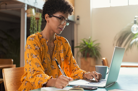 Focused college student sitting in cafeteria taking notes while using laptop. Young brazilian woman doing research for business at coffee shop. African american girl sitting in cafe using computer and writing notes.の写真素材