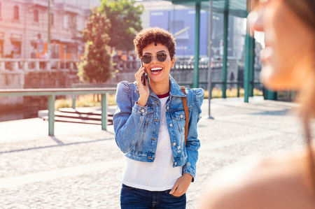 Cheerful young woman laughing while talking on mobile phone in the city centre. Excited brazilian girl walking while using smartphone on city streets. Happy african stylish girl enjoying journey while talking on cellphone.の写真素材