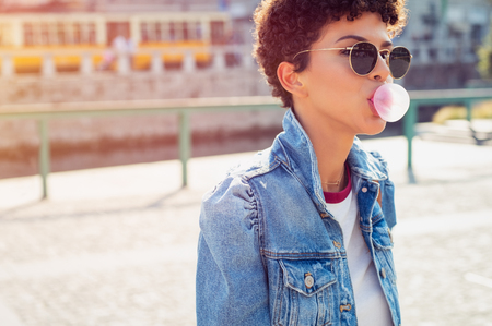 Young beautiful african woman in sunglasses with curly hair puffs bubble of chewing gum in summer. Brazilian young woman inflates pink chewing gum. Stylish woman making bubblegum of chewing gum.の写真素材