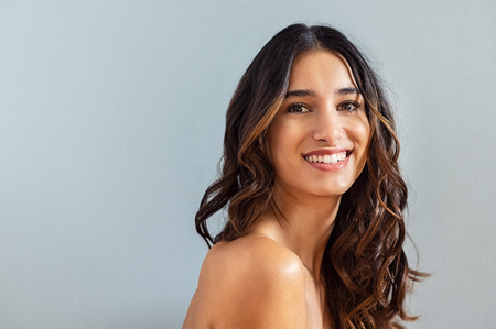 Smiling young woman looking at camera isolated on gray background. Beauty portrait of brunette girl with long curly hair looking at camera. Skin care treatmet and haircare concept at beauty spa.の写真素材