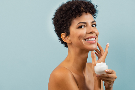 Beautiful young woman holding jar of skin cream for face. Happy brazilian woman applying anti aging moisturizer and looking at camera with copy space. Smiling african beauty applying face moisturizing cream isolated on blue background.の写真素材