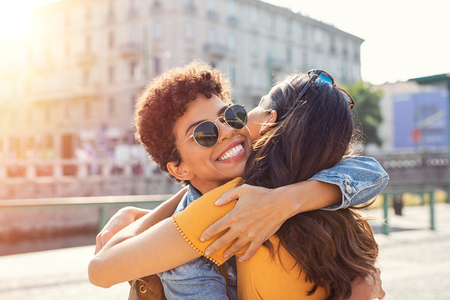 Happy meeting of two friends hugging in the street. Smiling girls friends laughing and hugging in the city centre. Multiethnic young women embrace each others after long time they have been distant.の写真素材
