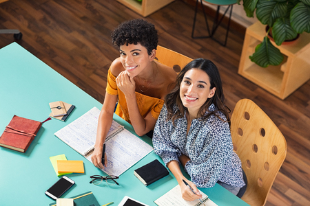 Top view of young women looking at camera while studying at library. Team of beautiful college students preparing for university exam. High angle view of smiling girl with her friend studying at cafeteria.の写真素材