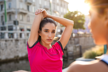 Young sporty woman wearing sportswear stretching arms outdoor. Active latin girl stretching arms over head during outdoor workout session with friend. Determined woman exercising on city street.の写真素材