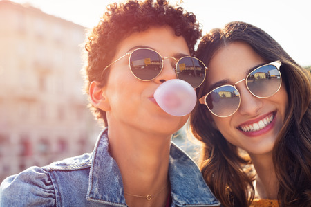 Portrait of brazilian woman with curly hair making bubble while latin girl smiling. Multiethnic friends enjoying vacation in city and looking at camera. Lesbian couple having fun while inflating chewing gum and making pink balloon.の写真素材