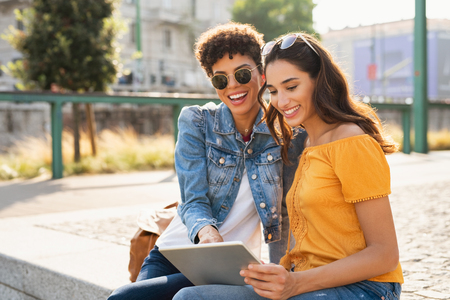 Two happy girls using digital tablet while sitting on stairs outdoor. Latin young woman looking for new sales on online store with her african friend. Multiethnic teens having fun while using laptop sitting on city street at sunset.の写真素材