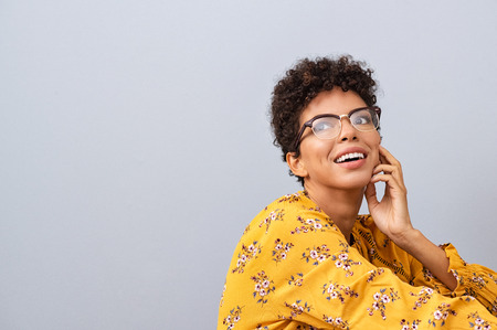 Happy thinking young woman looking away with fashion eyeglasses. African american female student thinking about future while isolated against grey wall. Brazilian with spectacles looking away and smiling.の写真素材