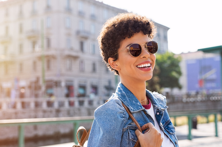 Portrait of young african woman in sunglasses and short curly hair looking away. Happy smiling brazilian girl enjoying summer on the street. Cheerful stylish black traveler enjoying vacation in the city.の写真素材