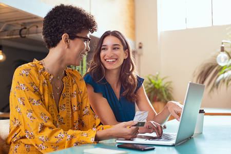 Happy woman making payment online using credit card. Cheerful friends discussing online shopping while purchasing products and paying bill amount using laptop and debit card. Two multiethnic girls sitting and relaxing at cafeteria.の写真素材