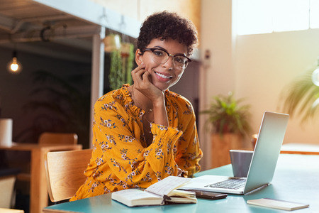 Happy brazilian girl with eyewear in coworking office with laptop looking at camera. Smiling african american student using computer at cafeteria. Successful young stylish business woman with glasses looking at camera.の写真素材