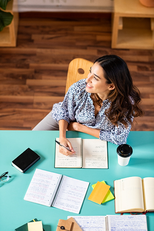 Smiling latin girl looking away while studying. Portrait of pensive college student looking away while studying alone at library. Young thinking woman taking notes at university.の写真素材