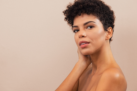 Young woman with natural skin isolated on background, looking at camera. Portrait of beauty black girl with curly hair touching her face skin. Mixed race lady with fresh daily makeup and copy space.の写真素材