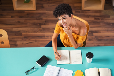Top view of beautiful smiling woman writing notes in book. Portrait of high school girl preparing for exam while sitting in cafeteria. High angle view of young african student writing notes while looking at camera.の写真素材