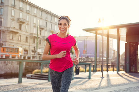 Smiling sporty woman running outdoors. Latin young woman jogging in the city street at susnset. Happy fitness girl wearing sportswear and exercising outdoor.の写真素材
