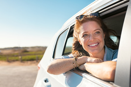 Smiling young woman peeking out of car window. Cheerful girl with freckles enjoying breeze from running car. Mature woman with red hair in the wind leans out of the car window and looking straight with copy space.の写真素材