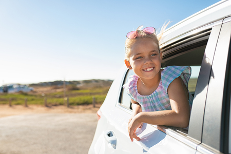 Cute blonde girl in the car looking through window. Smiling little girl having fun travelling by car and looking out. Happy smiling child sticking her head out of window and looking road trip.の写真素材