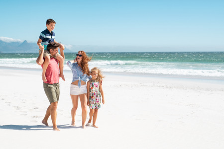 Smiling parents with children at beach. Cute son sitting on father shoulder with mother and sister walking while talking. Happy family with two children enjoying summer holiday at beach with copy space.の写真素材
