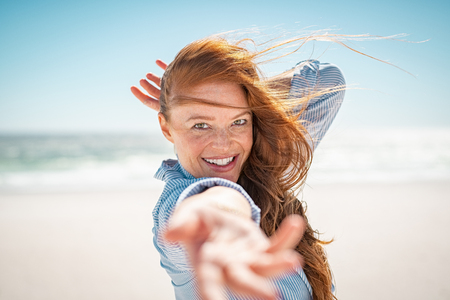 Cheerful young woman with red hair enjoying holiday at beach. Beautiful mature woman at sea during a windy day. Attractive girl smiling and looking at camera while feeling free at beach.の写真素材