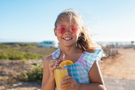 Cute little girl with glass of orange juice at outdoor park. Portrait of happy smiling child drinking pinapple juice outdoor and looking at camera.  Fashion girl wearing pink sunglasses holding glass with straw.の写真素材