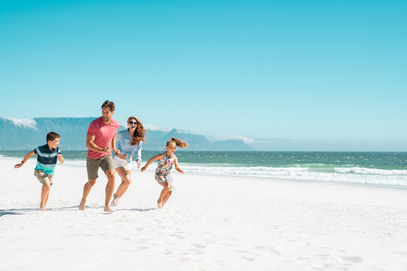 Happy young family running on the beach with copy space. Beautiful mother and laughing father enjoying summer vacation with son and daughter. Smiling parents with two children in casual having fun at seaside.の写真素材