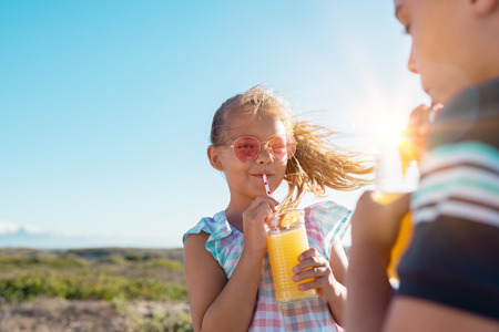 Portrait of little girl drinking orange juice in a glass with straw. Rear view of brother drinking with sister at outdoor park. Boy and cute girl with sunglasses suck from the straw a fresh pineapple juice outdoor with copy space.の写真素材