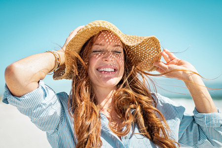 Closeup face of mature woman wearing straw hat enjoying the sun at beach. Happy young woman smiling during summer vacation at sea. Portrait of beautiful lady relaxing at beach while holding large brim for the wind.の写真素材