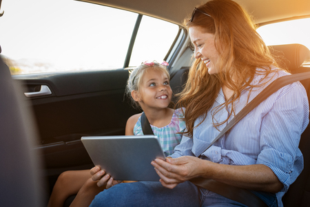 Portrait of smiling mother and happy little girl using digital tablet together in car. Mature woman showing videos to daughter in digital tablet while sitting in a taxi. Cute child learning new things with mother from computer.の写真素材