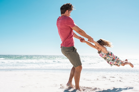 Father and daughter playing together on the beach with copy space. Happy dad and cute smiling little girl playing at seaside. Young daughter having fun at beach with ring around the rosie.の写真素材