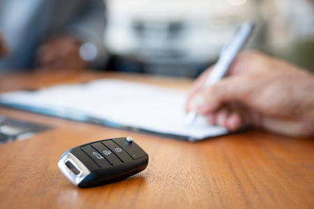 Car keys on desk with man signing purchase documents in background. Closeup of black modern car keys while hand complete the insurance policy or rental documents. Guy buying new car at dealership.の写真素材