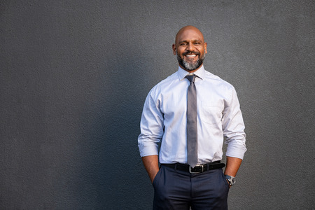 Portrait of successful black businessman standing against grey wall. Handsome senior entrepreneur wearing blue tie and formal clothing smiling and looking at camera. Mature happy man with hands in pocket isolated on grey background with copy space.の写真素材