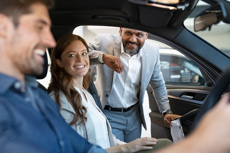 African salesman showing new car to young couple in showroom. Happy man and woman choosing auto in car dealership while cardealer explaining the new technology. Smiling beautiful couple test the comfort of their new suv.の写真素材
