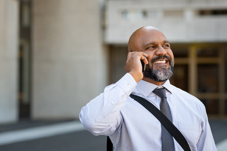 Mature african businessman talking on smartphone with modern building in background. Happy professional business man using cell phone outside, in city center. Portrait of senior man with beard talking on the phone and smiling with copy space.の写真素材