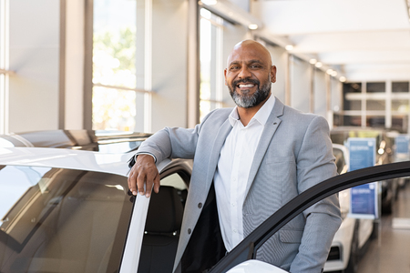 African salesman standing in car showroom near opened door of luxury car. Happy businessman buying his new suv in car salon. Smiling mature man choosing his new car.の写真素材