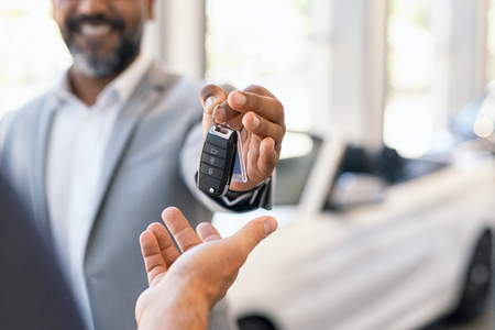 Closeup hand of cardealer giving new car key to customer. Detail of salesman hand giving keys to a client at showroom. Man's hand receiving car keys from african agent in a auto dealership with copy space.の写真素材