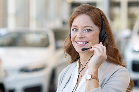 Beautiful female customer support wearing headset and looking at camera. Portrait of smiling woman customer service representative working with cars in background. Young call center operator at car rental office.