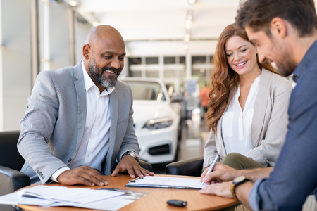 Cheerful couple and african salesman completing documents after buying new car. Young man signing car contract while sitting with woman and cardealer at showroom. Happy guy writing paperwork while purchasing and insurance his new automobile.の写真素材