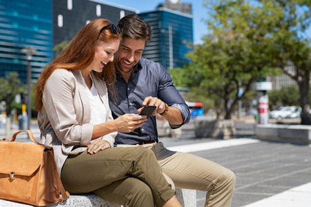Happy young couple using smartphone together outdoor in a modern square. Smiling mature couple using cellphone and social media while relaxing on a bench in a street. Young man and beautiful woman in love using mobile phone.の写真素材