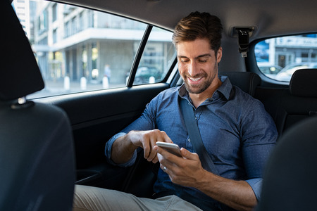 Happy smiling businessman man typing message on phone while sitting in a taxi. Young businessman in formal clothing using smartphone while sitting on back seat in car. Cheerful guy messaging wirth cellphone.の写真素材