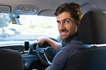 Portrait of handsome man driving car to work. Smiling casual businessman turning behind while sitting on driver seat. Satisfied young man driving his new car while looking at camera.の写真素材