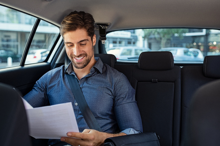 Business man looking at documents with concentration in his car. Young happy businessman reading contract on car back seat and smiling. Successful entrepreneur reading paperwork documents while sitting in taxi.の写真素材